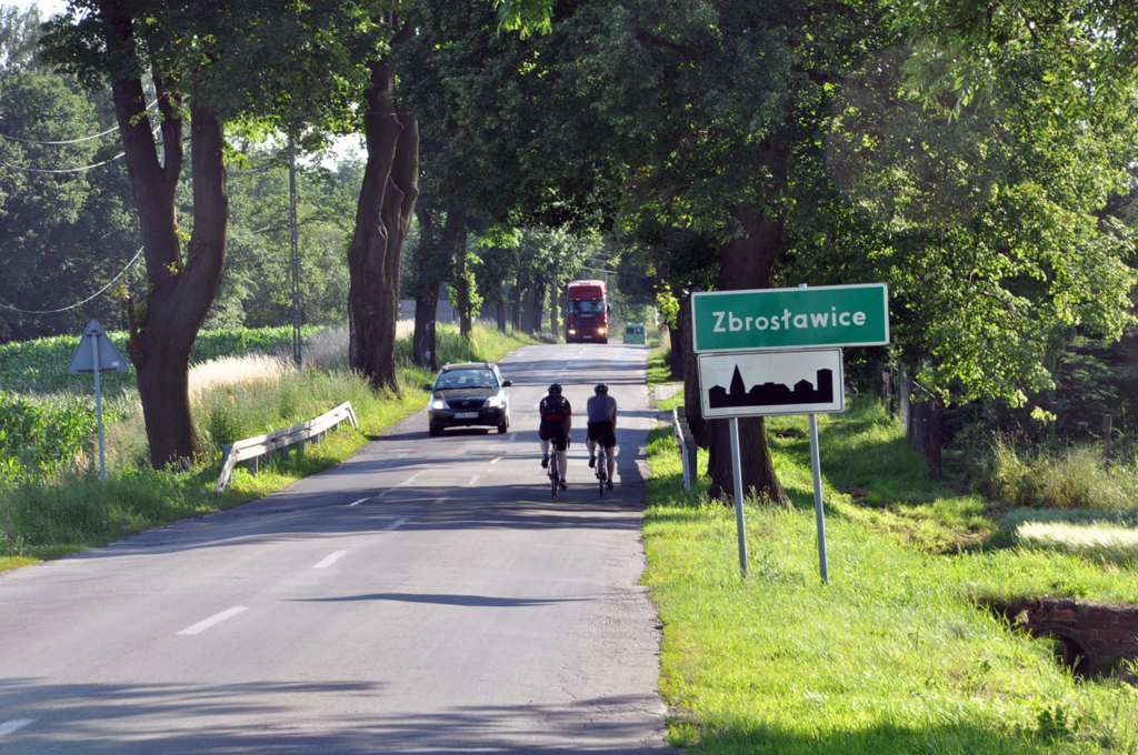 Droga, samochody i rowerzyści. Na poboczu znak dwa znaki - Zbrosławice oraz wjazd w teren zabudowany. 